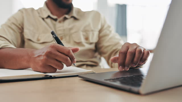 Hands, laptop and a man writing in a notebook while sitting at his desk, working on a schedule in the office. Computer, planning and administration with a male employee at work to write a diary note