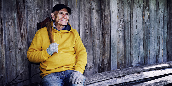 Senior lumberjack with old iron axe resting against wooden rustic barn wall. Mature woodman sitting outdoor.