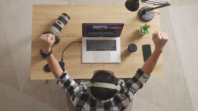 Top View Of A Male Editor With Headphone Celebrating Succeed Using A Laptop Next To The Camera Editing The Video In The Workspace At Home