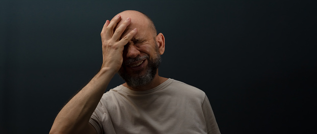 Sad man suffering from headache or migraine. Stressed guy with painful facial expression feeling terrible weakness or depression. Isolated on dark background.