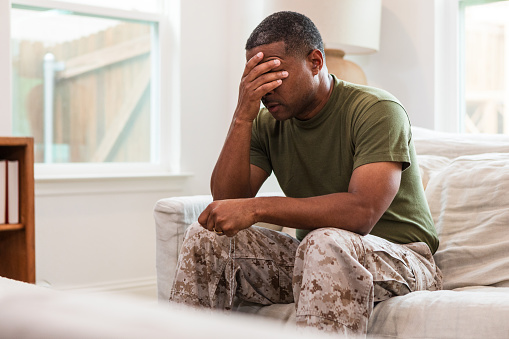 The distraught mature adult soldier covers his eyes with his hand as he sits alone in his living room.