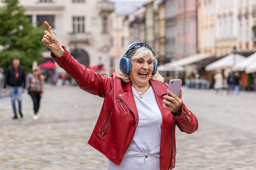 Happy overjoyed senior woman in wireless headphones listening favorite energetic music, celebrating win. Elderly grandmother tourist traveler walking in urban city street. Town lifestyles outdoors