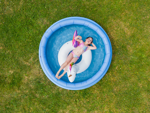 A little girl swims on an inflatable unicorn ring in an inflatable pool in the garden in summer, aerial view. High quality photo