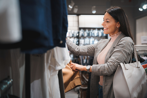 Smiling young female choosing new clothes in shopping center. Modern stylish woman looking for a new dress in a shopping mall.