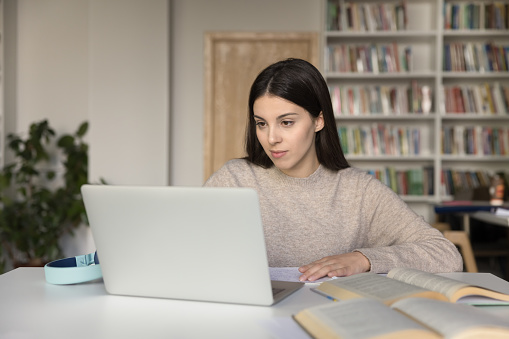 Attractive student girl sits at desk in library prepare for university exams or high school admission using laptop looks focused, makes exercise, browse information, do task. E-learning, modern tech