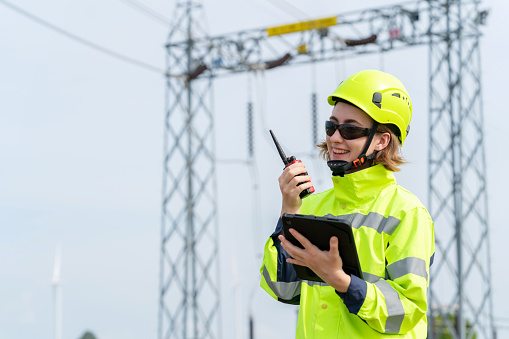 Engineer utilizing tablet and radio communication to coordinate with manager at power plant site at a wind turbine location. Focus on emphasis on technology, environmental conservation, and renewable energy. Confident demeanor showcases their expertise in managing power plant systems.