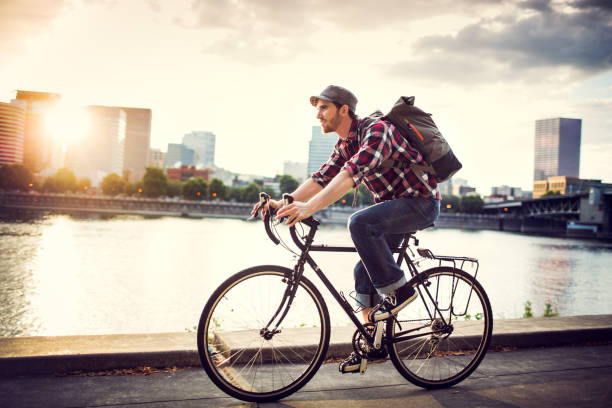 Bike Commuter in Portland Oregon A young man commuting in an urban city environment on his street bicycle, a waterproof bike bag on his back. This is the Eastbank Esplanade in Portland, Oregon, that follows along the Willamette river. Downtown is visible across the water, the sun shining brightly between the buildings. Horizontal with copy space. In-camera lens flare. hipster bike stock pictures, royalty-free photos & images