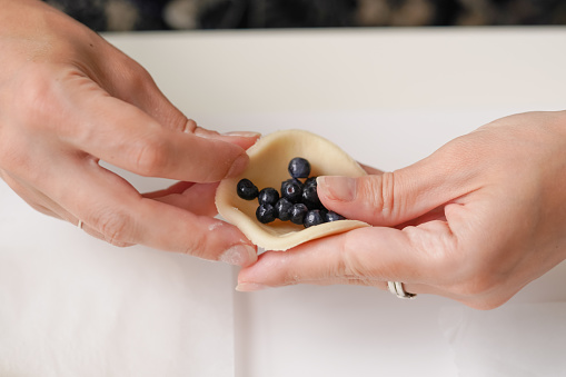 Women's hands make Ukrainian varenyki with blueberries on a light background. A close-up of the preparation of a traditional dessert made of dough and berries