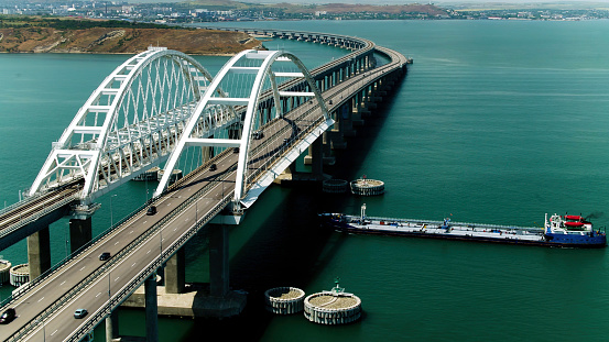 A ship passes under a large bridge with moving cars. Shot. Aerial view of a bridge and a ship with turquoise water, time lapse effect