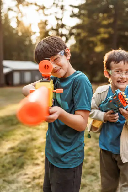 A Eurasian tween boy and his younger brother smile and take aim while having a playful nerf gun battle in the back yard of their home on a warm summer evening. Both boys are wearing protective eyewear.