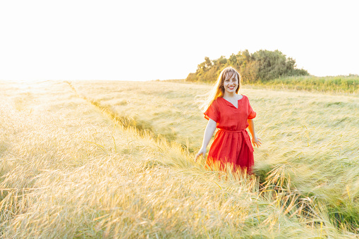 Young happy woman in red dress walking in wheat field on sunset. Breathe of freedom. Positive emotions feeling life, peace of mind. Mental health practice. Nature relaxation. Soft selective focus.