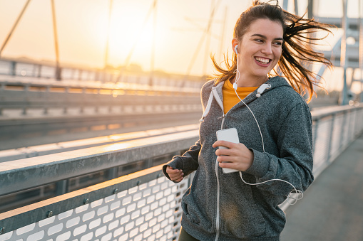 Cheerful sportswoman listening to music on earphones while jogging on a bridge at sunrise. Happy girl doing her preparation training for marathon.