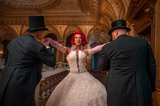 A beautiful elegant 1920s style redhead drag queen in a luxury stately home with handsome male chaperones