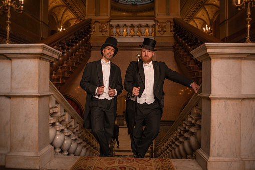 Two handsome 1920s style gentlemen wearing white tie and top hat on a luxury stately home staircase