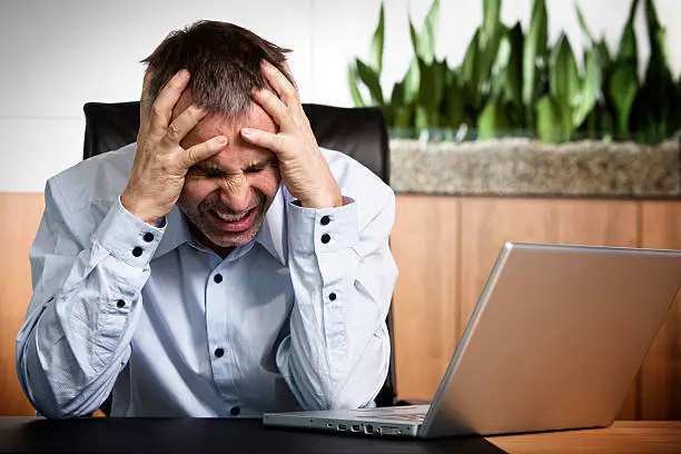 Frustrated overloaded senior businessman being furious about work and clinching his teeth, sitting at office desk in front of laptop.