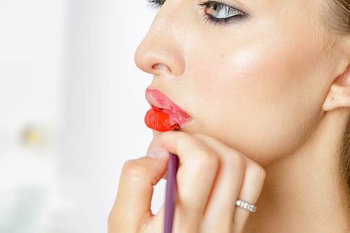 Close up of hand of visagiste applying lipstick on female lips. She is holding a brush. The young beautiful woman with red lip