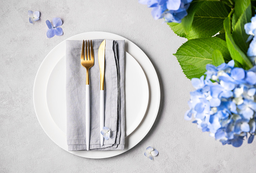 Festive table setting with blue hydrangea flowers and white plates on a light background. The concept of celebrating women's day, birthday or mother's day. Top view and copy space.