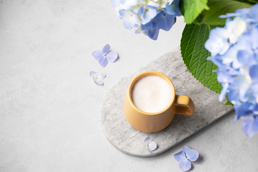 Spring bouquet with blue hydrangea flowers and a cup of cappuccino coffee in a yellow cup on a light background. The concept of a morning drink for breakfast. Top view and copy space.