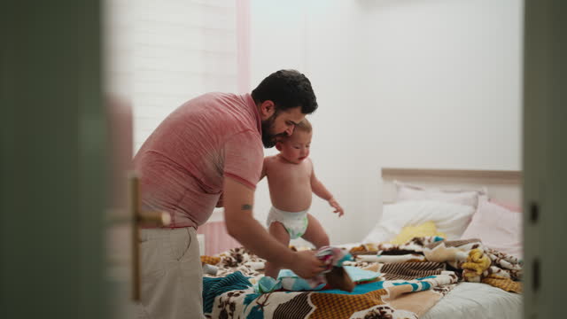 Father taking care of his small baby boy and changing his clothes and baby diaper in bed room at home