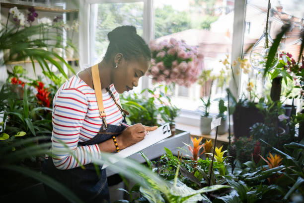 Side-view of female florist Black ethnicity taking inventory of plants at the flower shop
