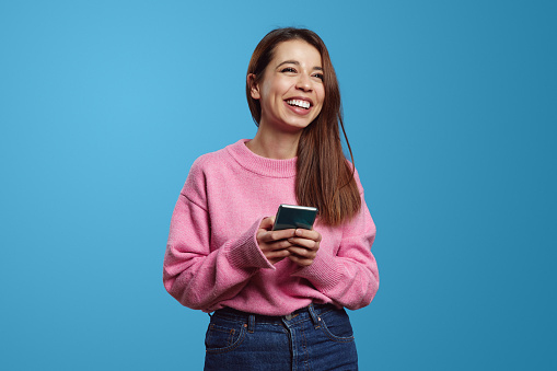 Excited latin girl looking away with a big smile while holding smartphone in hands, standing isolated over blue background