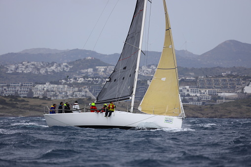 Bodrum,Mugla, Turkey. April 02,  2023: sailor team driving sail boat in motion, sailboat wheeling with water splashes, mountains and seascape on background. Sailboats sail in windy weather in the blue waters of the Aegean Sea, on the shores of the famous holiday destination Bodrum