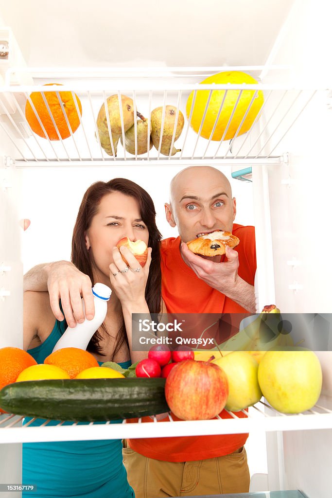 Couple eating cakes A couple eating cream cakes standing in the open door of the refrigerator full of healthy fresh fruit. Food Stock Photo