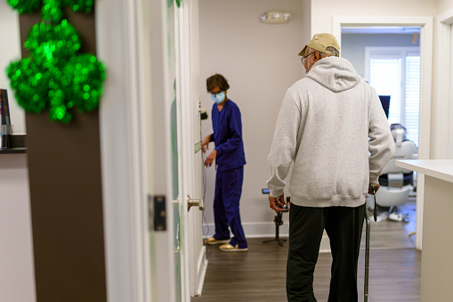 Senior, male patient of Indian descent walks, with a cane, to an exam room at a dental office, following a female medical professional.