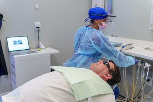 White, male patient lays on a dental chair while his multiracial, female dentist takes a pause during a check up appointment.