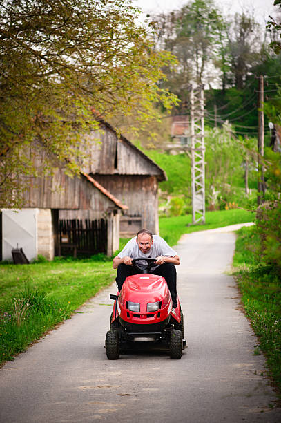 Cheerful gardener riding tractor mower on countryside road. Shallow DOF. stock photo