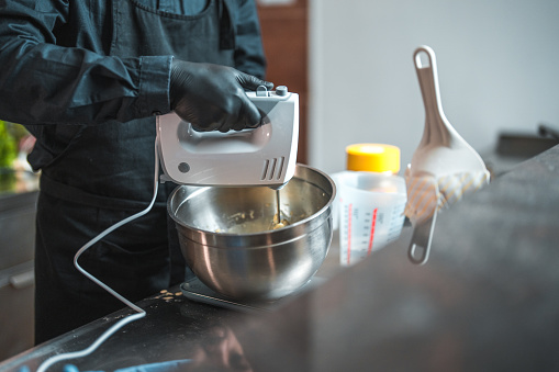 Male hands using black gloves and handling a hand mixer to blend ingredients. Chef takes charge of the mixing process, carefully combining ingredients in a metal bowl.