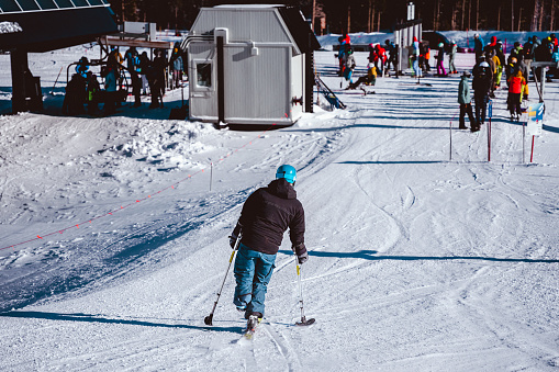 Rear view of an adaptive athlete using a single ski and handheld outriggers nearing the bottom of a ski run, with a queue of unrecognizable skiers and snowboarders waiting for the ski lift visible in the distance.