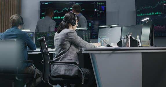 Diverse financial analysts monitor real-time stocks on computers. Team of businesspeople work in bank office. Big digital screens showing exchange market charts. Cryptocurrency trading and analytics.