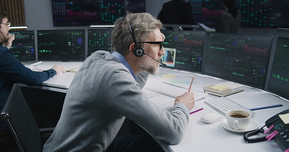 Mature male trader in headset works on multi-monitor computer with real-time stocks in office. Colleagues analyze exchange market charts on big digital screens. Cryptocurrency trading and analytics.