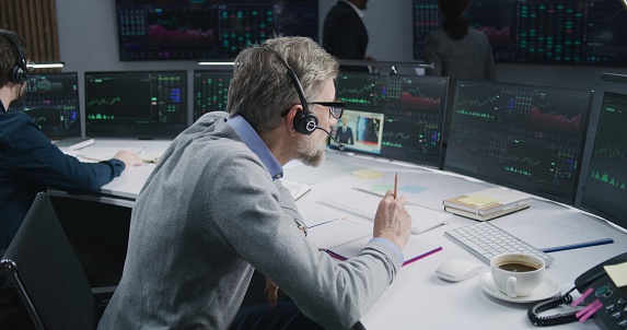 Mature male trader in headset works on multi-monitor computer with real-time stocks in office. Colleagues analyze exchange market charts on big digital screens. Cryptocurrency trading and analytics.