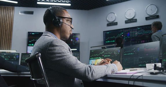 African American investor with pen in hand works at computer with displayed real-time stocks. Colleagues analyze exchange market charts on big screens at background. Concept of trading and investment.