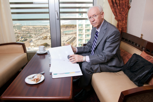 An elderly (in his 80's) business man wearing suit and tie sitting in a hotel's business lounge, looking at camera, as if disturbed in the middle of going over some papers after having coffee. The sea and a marina can be seen defocused in the background.