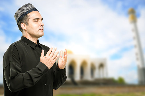 A Muslim man in Asia is praying, in the background of a mosque.