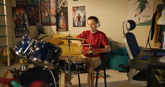 Young boy in headphones practices musician skills using drum kit at home.