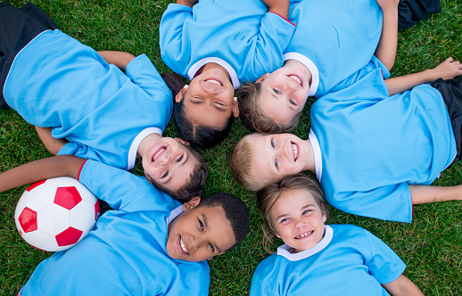 A small group of children lay in the grass with their heads together as they pose for a portrait after a soccer game.  They are each wearing a blue jersey and are smiling.