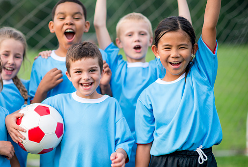 A small group of elementary aged children cheer after a soccer match as they celebrate their win.  They are each wearing a blue jersey and are smiling.