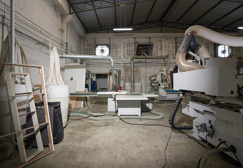 Atmosphere inside wooden furniture factory with piles of plywood and modern machinery. An exhaust fans are on lightweight concrete brick wall in the background.