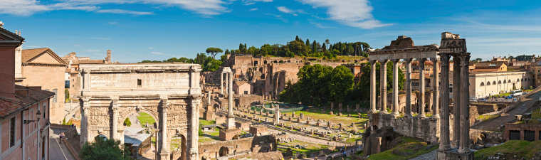 Warm late afternoon sunlight casting long shadows across the ancient temples and arches, palaces and churches of the Foro Romano, the iconic arcades of the Coliseum and the leafy foliage of Palatine Hill in the heart of Rome, Italy under panoramic summer skies. ProPhoto RGB profile for maximum color fidelity and gamut.