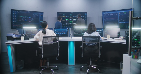 Female software engineer sits at computer with displayed blockchain network and surveillance maps. Technical support specialists work in monitoring room. Multiple big screens on the wall. Dolly shot.