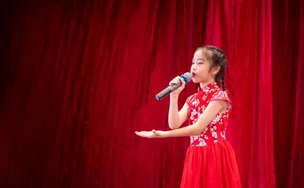 Photo of The Asian kid girl sing a song on stage at her school activity day, dress in Qipao style, red curtain background