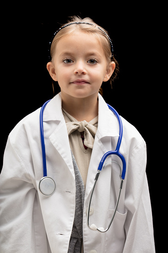 Small girl plays doctor, half-body portrait on black background. She wears an oversized white coat and stethoscope. This cute blonde girl might prefer to become a vet when she grows up