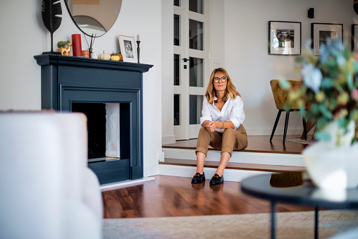 Full length of attractive blonde haired woman wearing white shirt and trousers while sitting on the floor in her modern home.