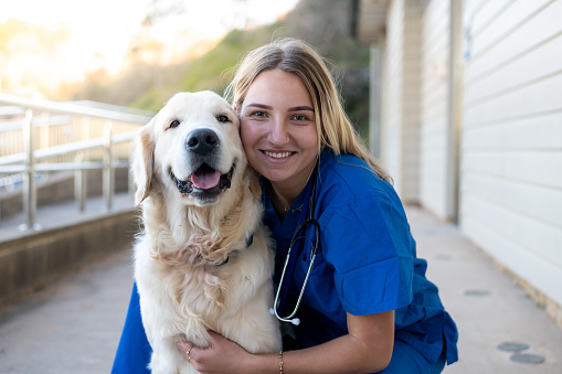 Female veterinarian hugging a beautiful golden retriever outside of an animal hospital. Looking at the camera and smiling. The doctor is wearing blue medical scrubs and a stethoscope around her neck.