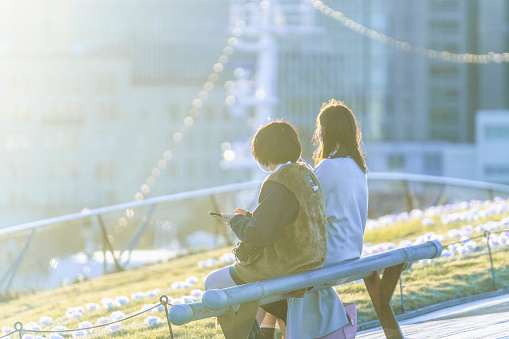 a serene moment in the bustling city of Yokohama, Japan. Two young women, each exuding contemporary style and confidence, are seen enjoying a leisurely afternoon on a bench. The woman on the left, dressed in a trendy ensemble, is engrossed in her phone, while her companion, dressed in a more traditional and elegant outfit, gazes thoughtfully at the cityscape. In the background, two other women are seen walking along a bridge, deep in conversation. The panoramic view of the city skyline, adorned with twinkling lights, provides a stunning backdrop. The image beautifully juxtaposes the tranquility of the park with the vibrancy of city life, encapsulating a shared moment of relaxation and connection.