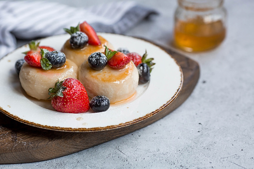 Cottage cheese pancakes, Russian cheesecakes with fresh blueberries, strawberries, honey and mint leaves on a stylish wooden board on a gray concrete table. Natural products. Healthy and delicious breakfast.